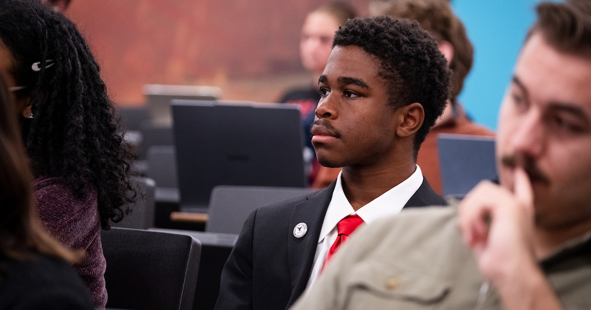 a young man with short, curly hair sits in a classroom while dressed in a black suit and red tie