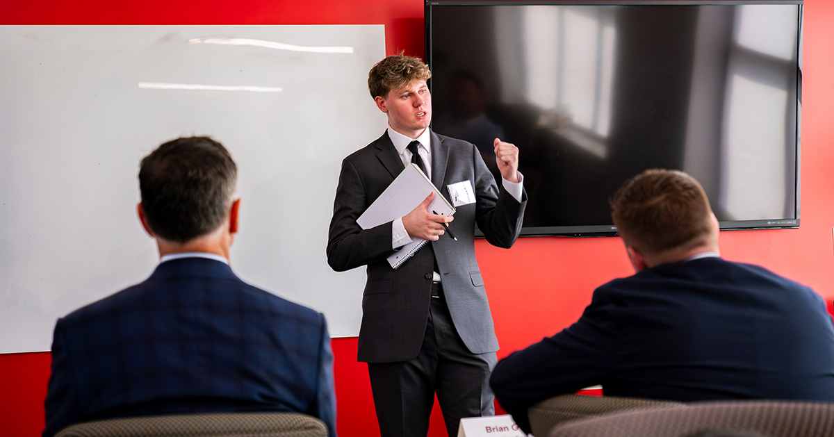 a young man dress in a suit and tie presents to a room of professionals with a whiteboard and large tv behind him on a red wall