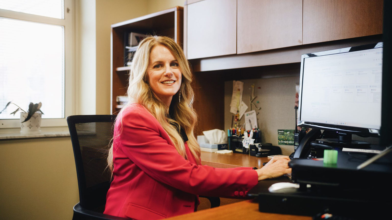 Amy Korus Ball State Alumna-Female in red suit coat with long blonde hair sitting at a desk with a computer monitor in front of her