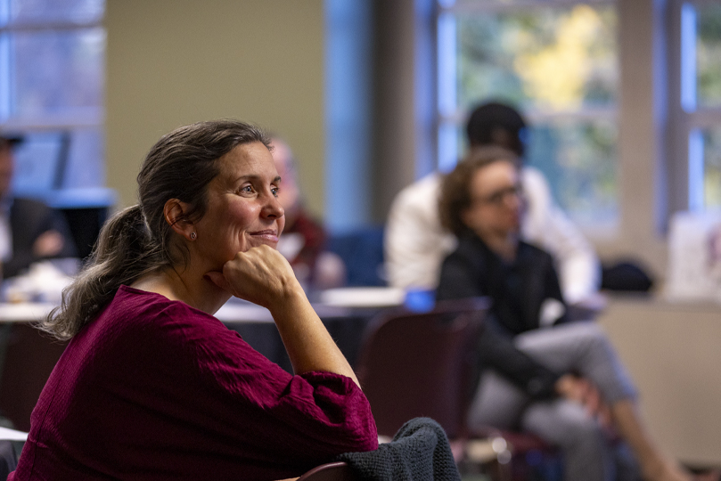 Middle aged woman with a long sleep maroon shirt sitting in a chair with her arm under her chin smiling listening to someone talk