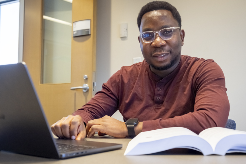 male student with glasses and a dark orange long sleeve shirt looking at the camera with a book and a laptop in front of him
