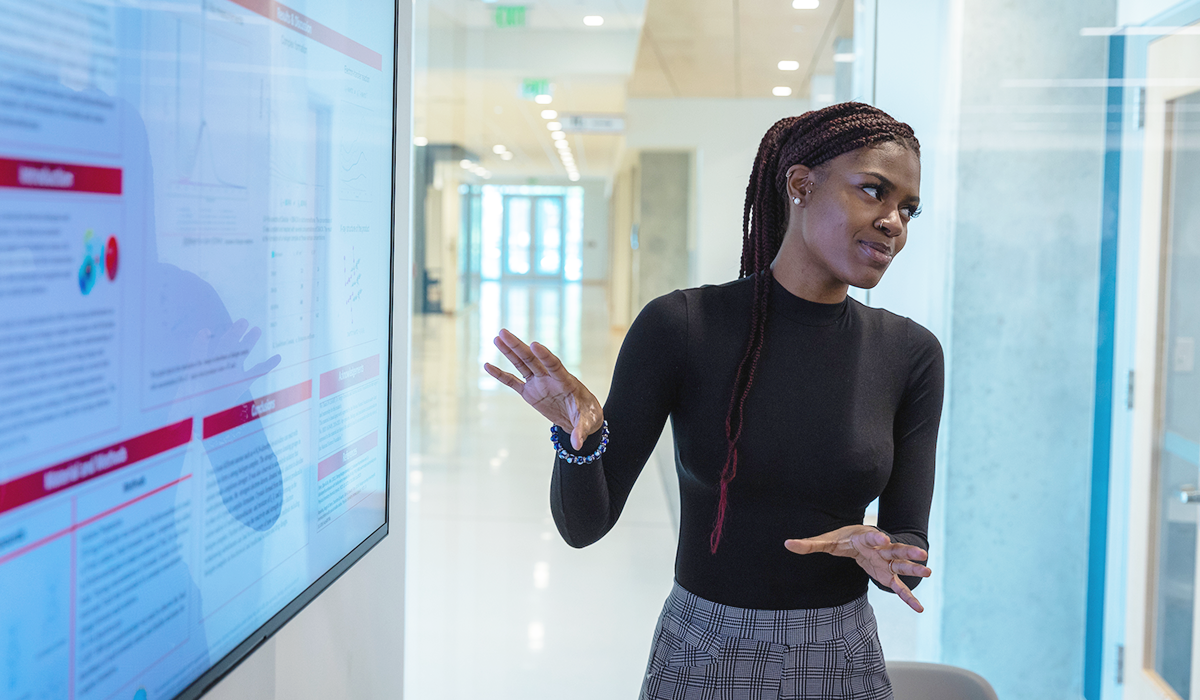 A business analytics student gesturing while speaking near a digital screen in a business classroom in Muncie, Indiana