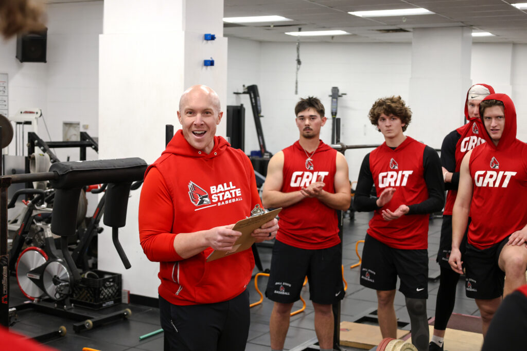 Bill Zenisek-man on the far left in red sweatshirt holding a clipboard, four young male students listening to bill speak. Located in a training room with workout equipment in the background