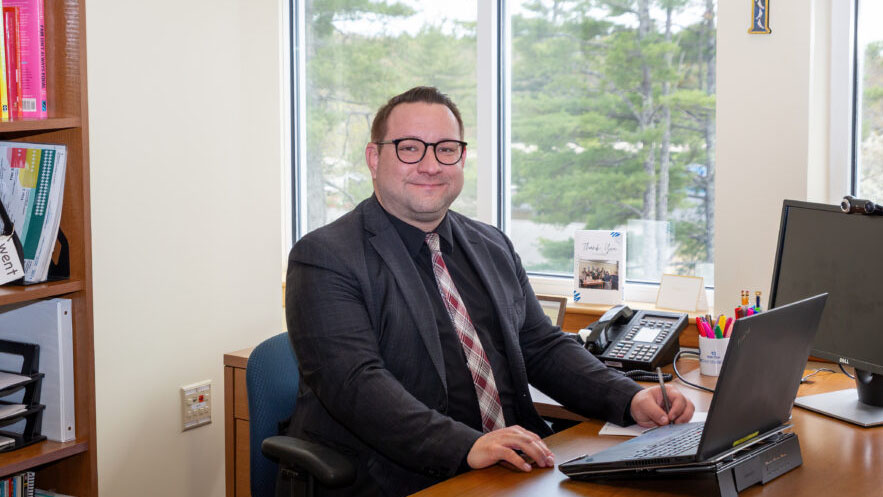 Stephen Marasitz, man in a black suit and tie sitting at a computer with a laptop open on the desk looking directly at the camera. Sitting in an office space with a bookcase to the left and windows behind Stephen at the desk.
