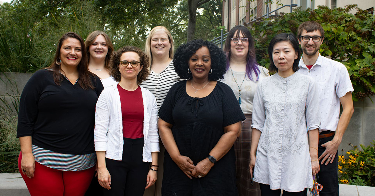 Eight faculty and staff members pose together outdoors in a landscaped campus courtyard.