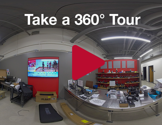 UMS Equipment Checkout with a large desk and several containers of telecommunications equipment and a screen on the wall.Click on the photo to take a 360° tour of the UMS Equipment Checkout.