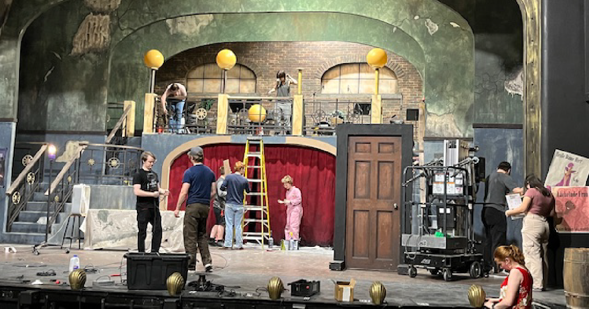 Several people working on a stage set that has green walls, a large wooden door, and a red curtain.