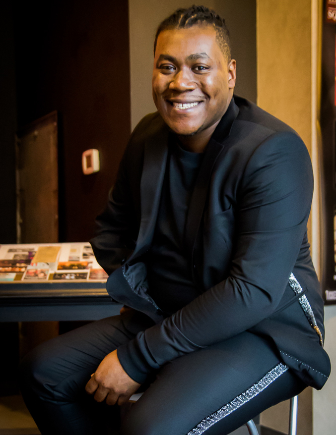 Photo of Emil Thomas. A Black/African American male, wearing a black button up shirt with a black suit.