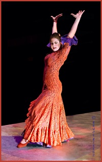A white women with brown hair. She is wearing a orange dress with purple on the inside of the sleeves. She is leaning back with her arms thrown up in the air.