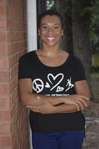 A Black/African American female wearing a black T-shirt with blue jeans. She is leaning up against a brick wall.
