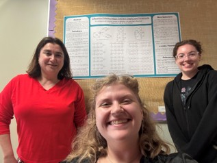 Three smiling people posing together indoors, with an academic poster displayed on the wall behind them.