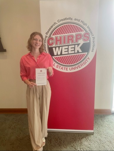 Olivia Glaeser holds an award or certificate beside a Ball State CHIRPS Week banner.