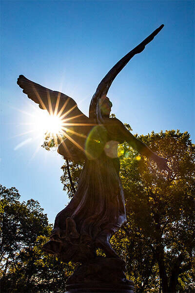 Ball States Beneficence Statue a bronze sculpture of an angel with outstretched wings silhouetted against a bright sunny sky 