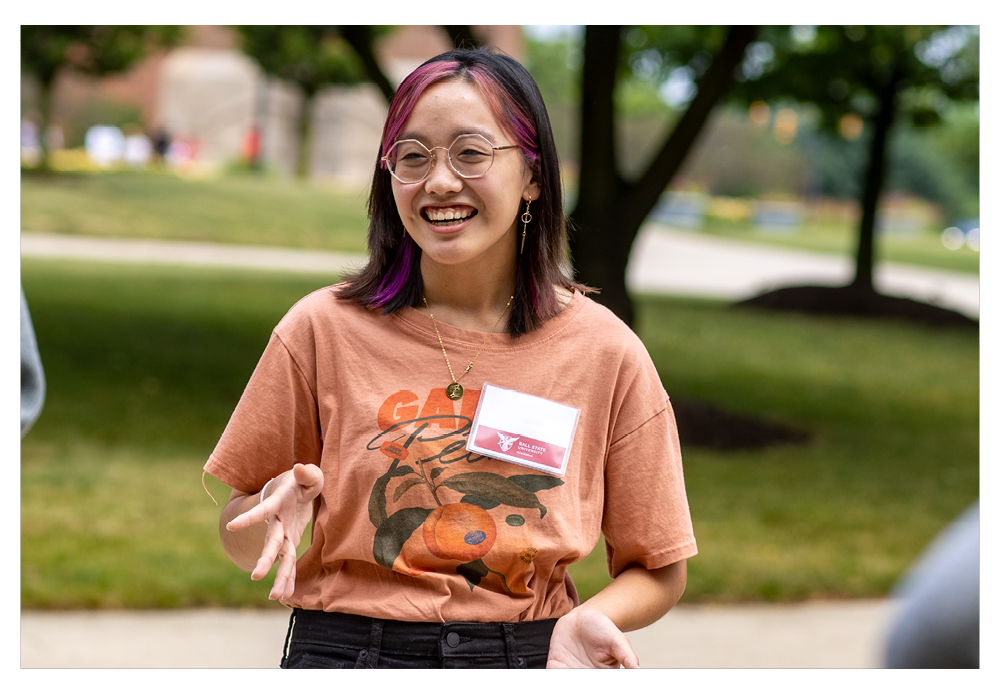 Female student with long brown hair with pink coloring, in orange shirt and orientation name tag, campus green and tree in the background