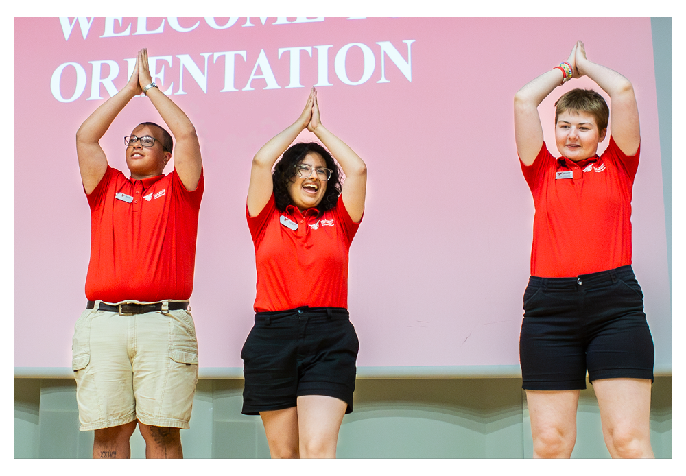 Three orientation leaders one on the left tall glasses and holding hands over head like an arrow, the middle female student holding hand above head and student on the far right holding their hands above their head