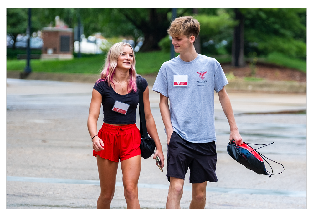 Two students walking one female student on the left with red shorts and a blue shirt with blonde hair, male student on the right with grey shirt and blue shorts. Walking on the sidewalk on campus