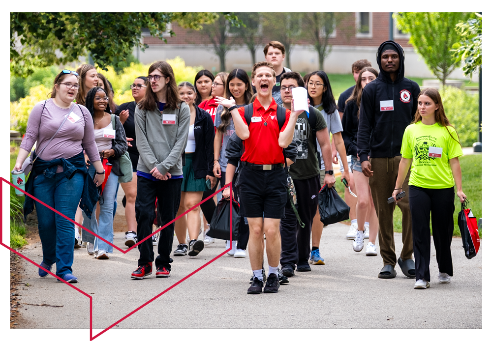 Group of orientation students walking through campus. A large group of about 20 students with the orientation leader in the front in a red shirt smiling and laughing