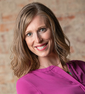 Smiling woman wearing a magenta blouse, posing in front of a light brick background.