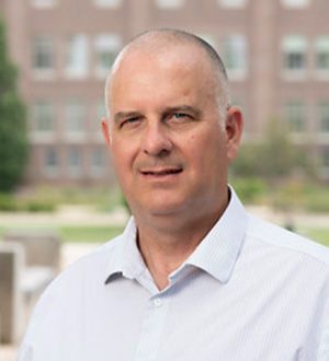 Man wearing a light blue button-down shirt, posing outdoors with campus buildings softly blurred in the background.