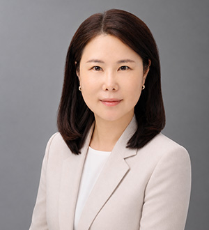 Professional headshot of a woman wearing a light-colored blazer against a gray studio background.