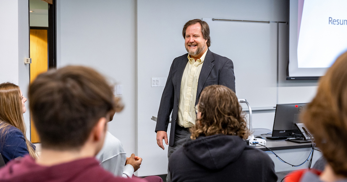 A man wearing a black blazer smiles while standing at the front of a classroom while looking out at students