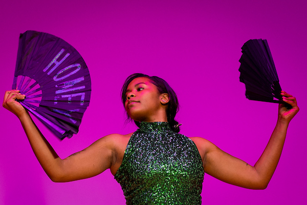 A woman is a aparky green halter dress dances with a set of fans against a bright purple backdrop.