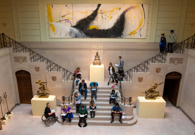 Lobby of the museum with people gathered on the grand staircase in the center. The staircase is bracketed by podiums displaying sculpture and a large painting is hung over the landing where the staircase splits into two separate sets of stairs to the left and right.
