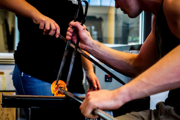 One person in a black shirt and jeans helps another person holding a glowing glass orb being held with metal forcepts.