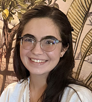 Gabriela Ramonell smiles for a portrait wearing glasses in front of a botanical-patterned background.
