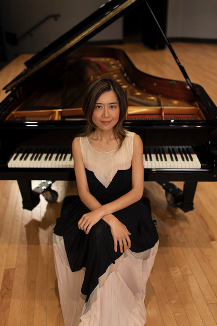 Jasmin Arakawa wears a black and white dress while sitting before a black grand piano on a polished wood stage.