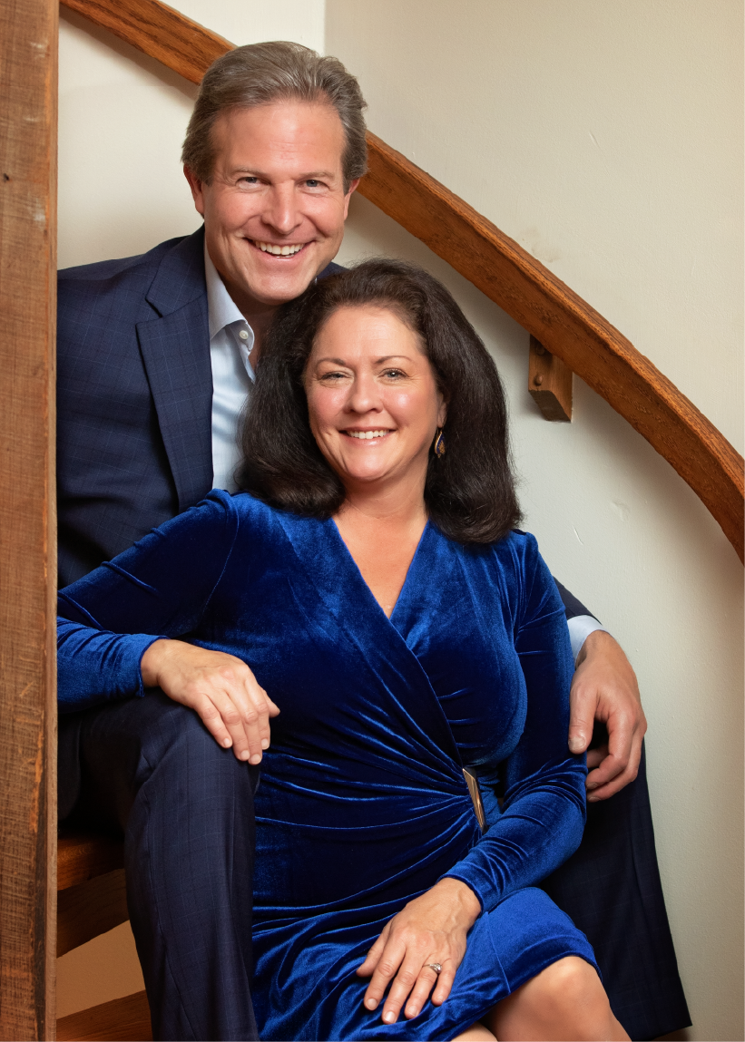 Dr. Daniel Narducci sits in a dark navy suit with his wife, Heather Narducci, in a white stairwell with a brown wooden railing. Heather wears a royal blue velvet dress.
