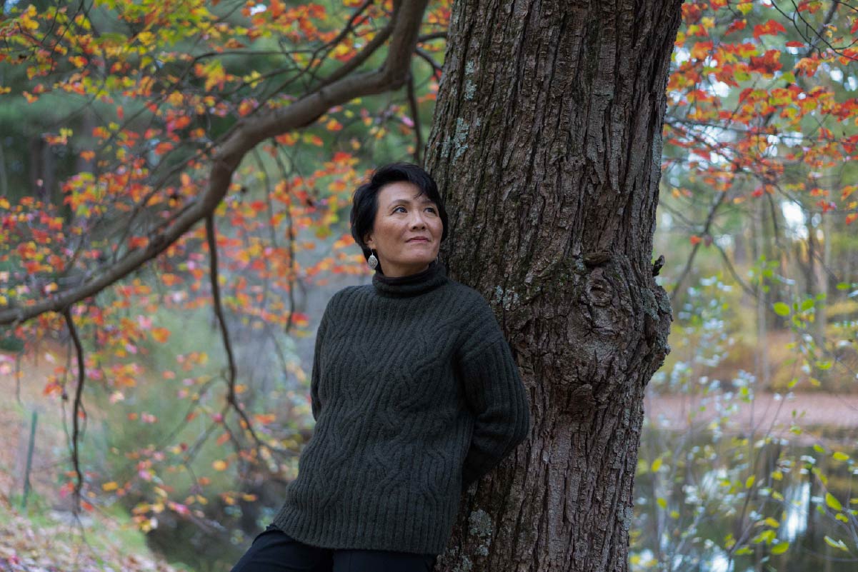 Mia Chung, pianist, leans against a tree with autumn colored leaves in a chunky gray sweater.