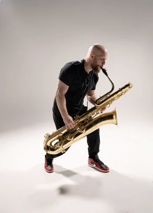 Dr. Nick Zoulek plays the saxophone in an all black outfit in front of a white backdrop.