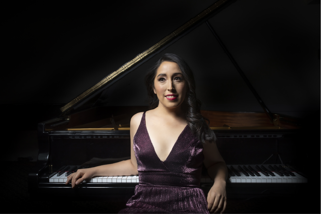 Priscila Navarro sits at a grand black piano wearing a sparkly dark purple dress