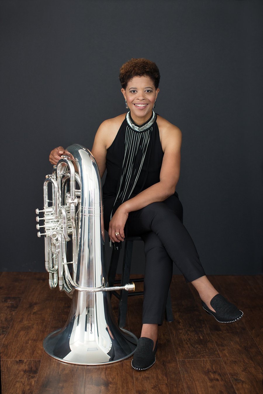Velvet Brown, musician, sits before a dark gray background wearing all black with metallic silver accents holding a silver tuba on her left.