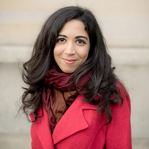 Woman posed in front of a white brick wall. She is wearing a red coat, and her long dark hair is down.