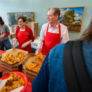 President and Mrs Mearns serving pizza with red aprons on from boxes to students