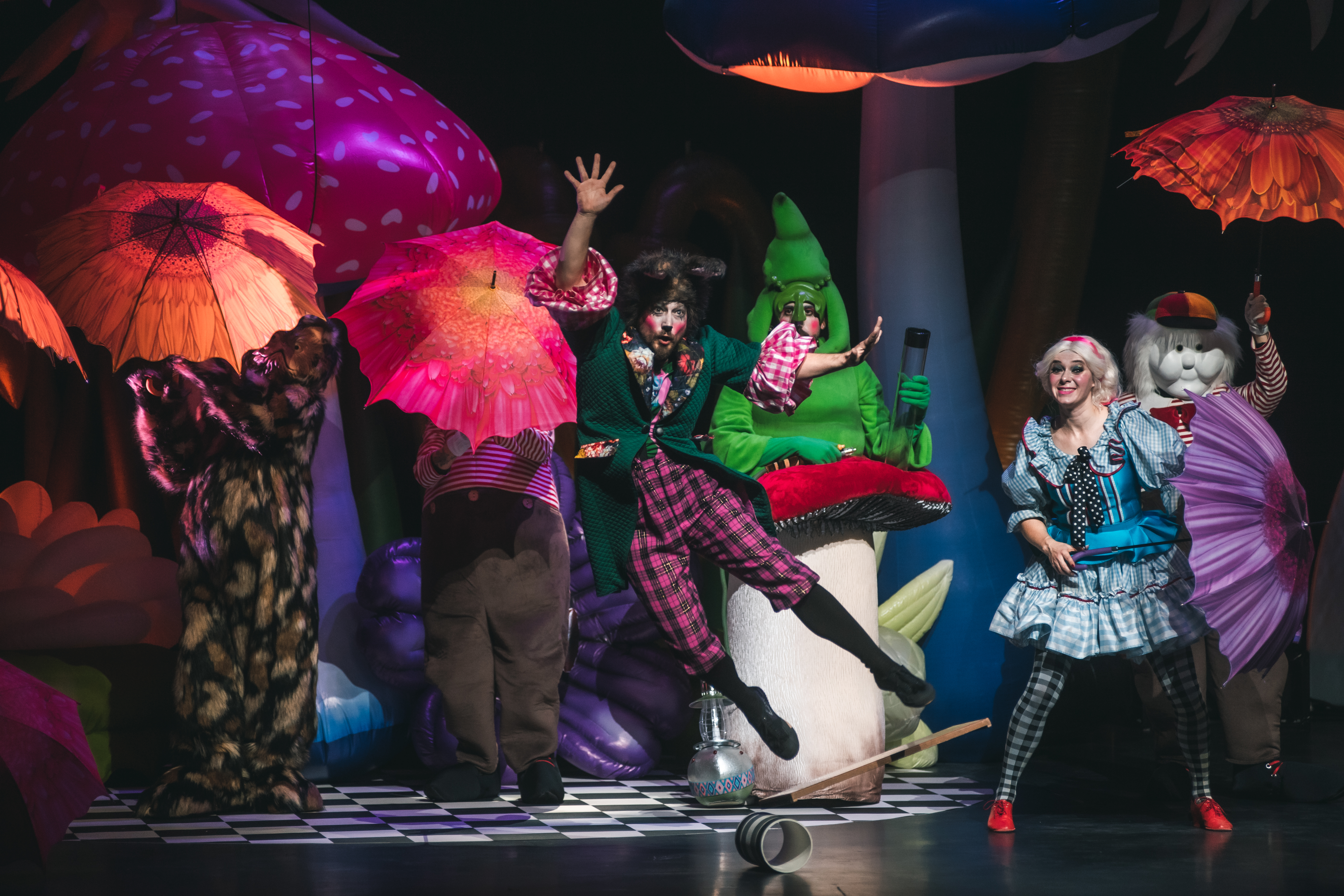 performers dance onstage in whimsical outfits in front of a background of colorful parasols