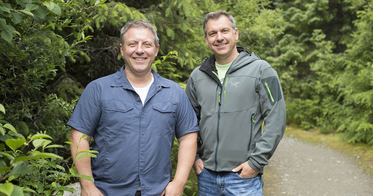  Wild Kratts brothers Chris and Martin Kratt smile pose with their hands in their pockets before green foliage and a gravel driveway.