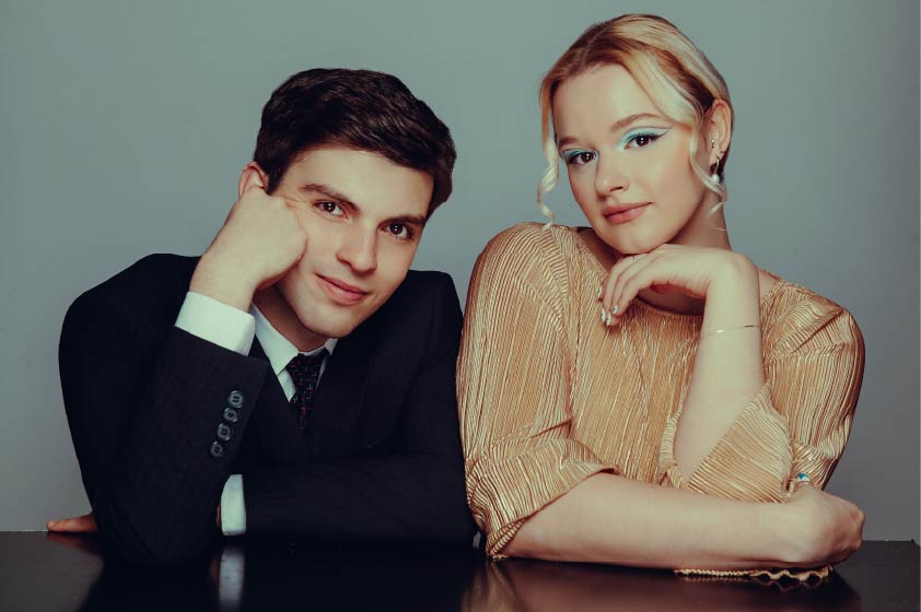 David Morino and Kate Kortum pose together before a slate blue background dressed in jazzy performance attire.