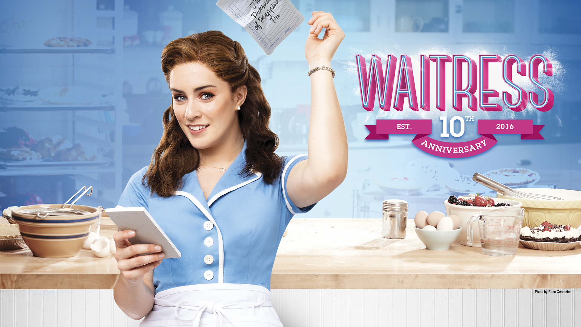 A woman waitress holding a notepad stands before a counter laid with various baking ingredients and pies, tossing a sheet to the side. She is accompanied by the Waitress logo. Image text reads, "Waitress 10th Anniversary – established 2016."