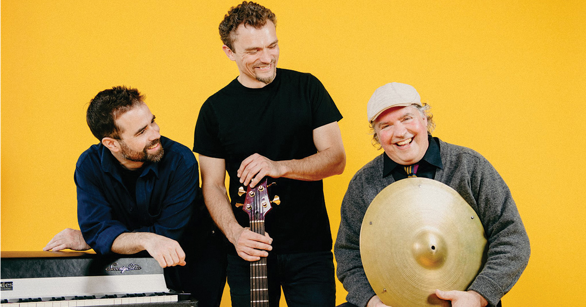 Three musicians smile at the camera posing with a keyboard bass guitar and cymbal against a yellow backdrop 