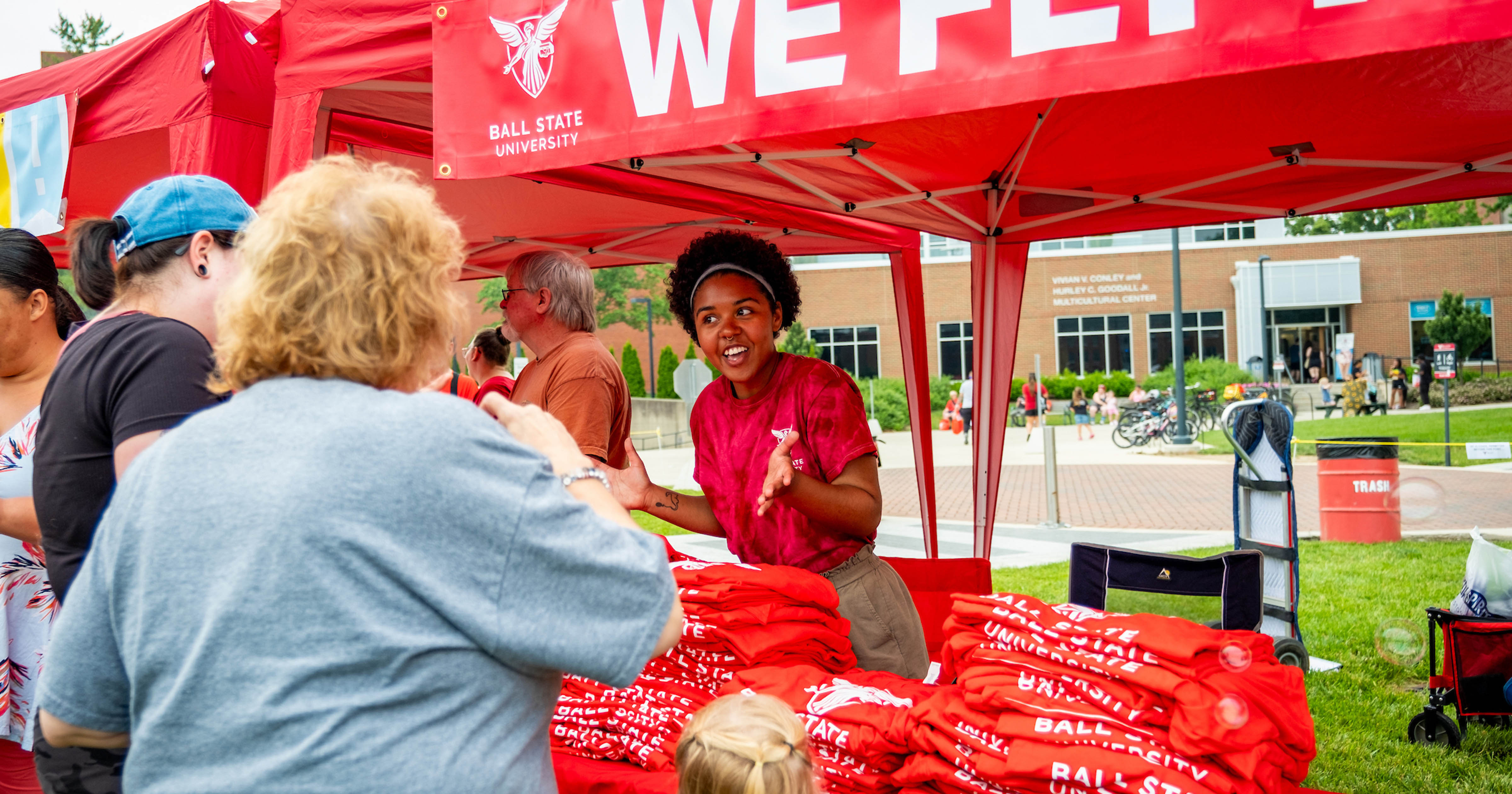 A Ball State student talks with community members at a red We Fly Fest tent filled with Ball State T-shirts and giveaways during an outdoor campus event.