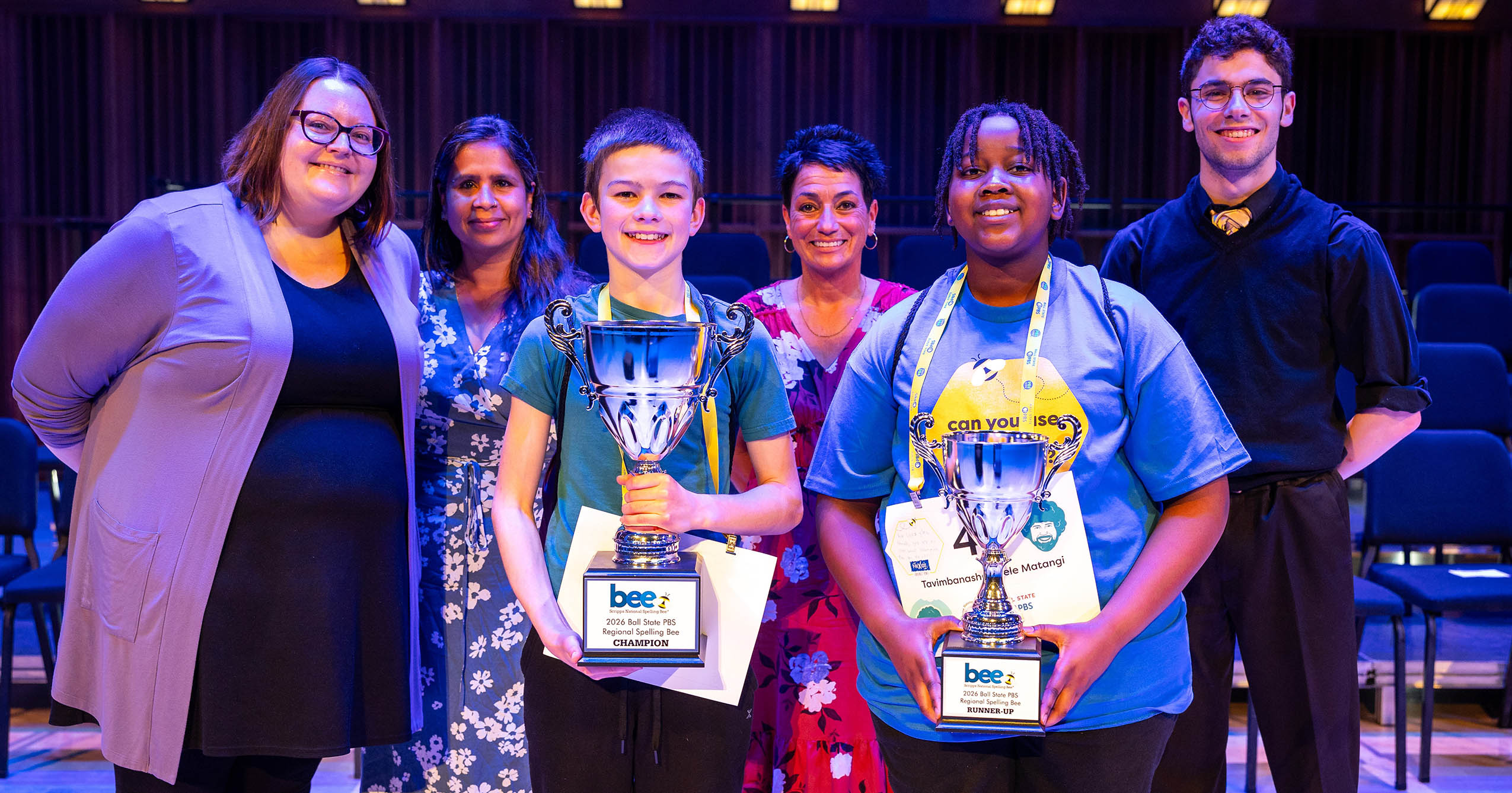 Leo Furlong and Tavimbana Anele Matani hold their spelling bee trophies onstage with event officials at the 2026 Ball State PBS Regional Spelling Bee.
