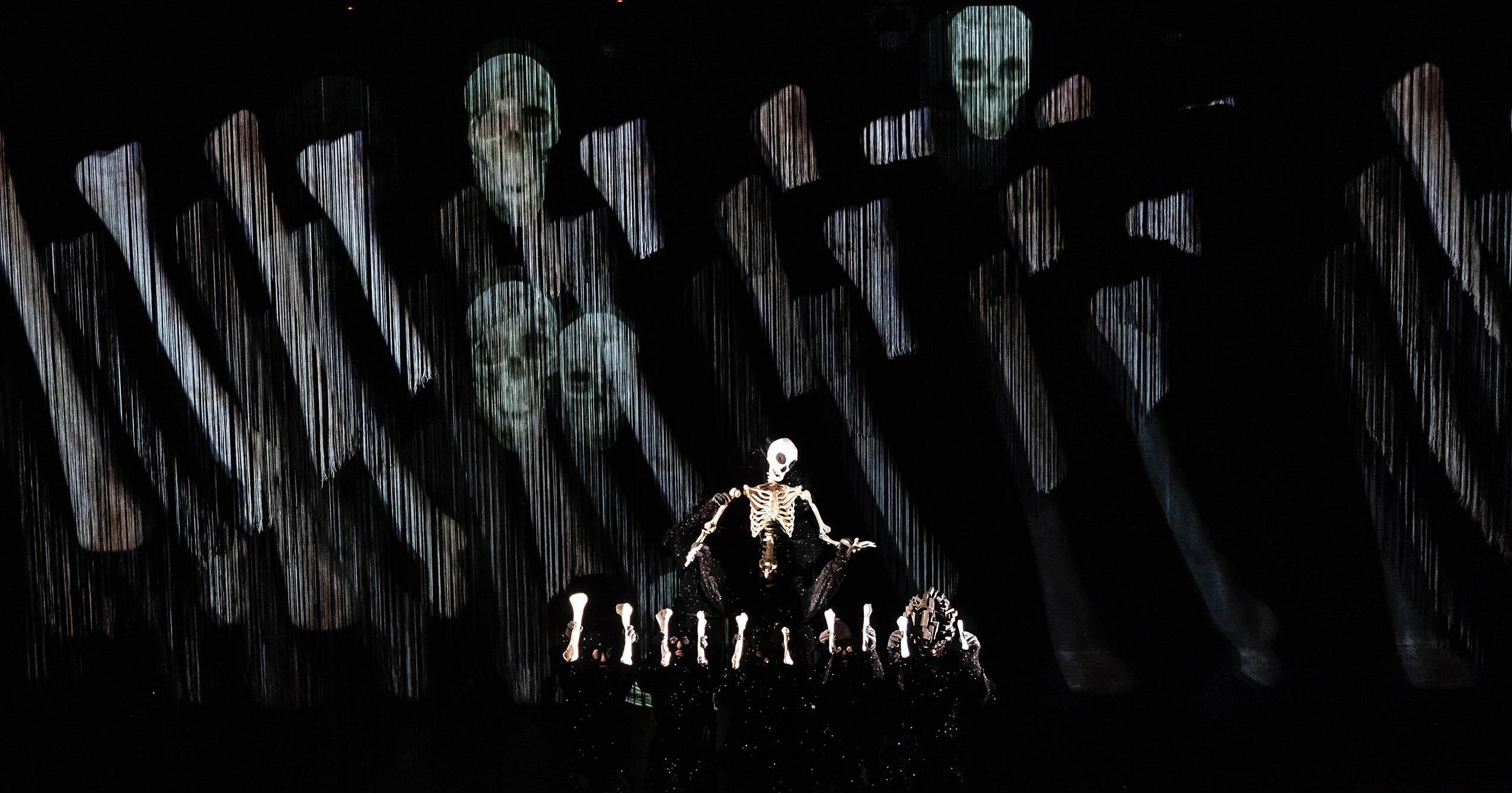 A skeletal puppet rises above performers holding illuminated bones, with ghostly faces and skeletal imagery projected onto a dark backdrop during Wakka Wakka Productions’ Dead as a Dodo.