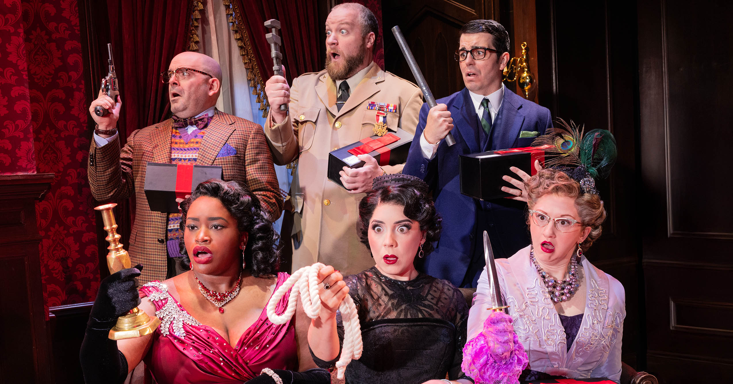 Cast members from the Broadway production of Clue pose onstage holding several of the mystery’s familiar weapons, including a revolver, wrench, lead pipe, candlestick, rope, and knife.