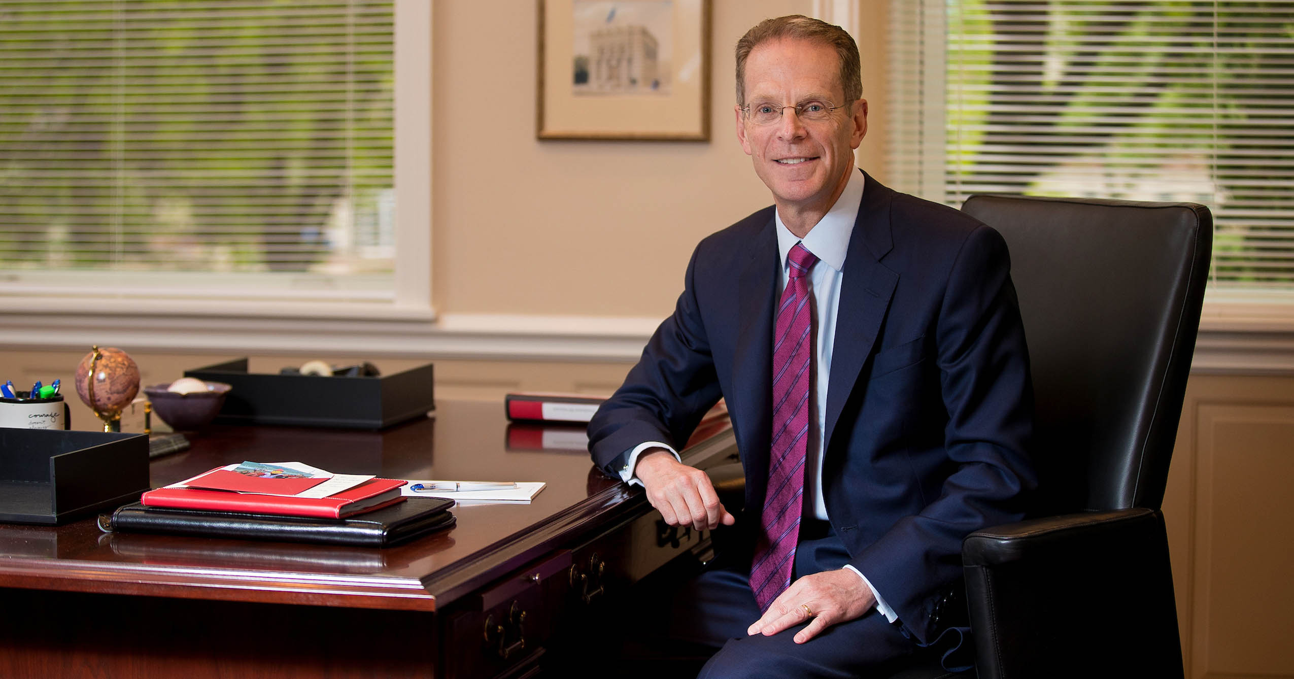 Ball State University President Geoffrey S. Mearns is seated in a black office chair beside a polished wooden desk. He is wearing a navy suit, light blue shirt, and red patterned tie. The office setting includes framed artwork on the wall, large windows with blinds, and desk items including folders, papers, pens, and a small globe.