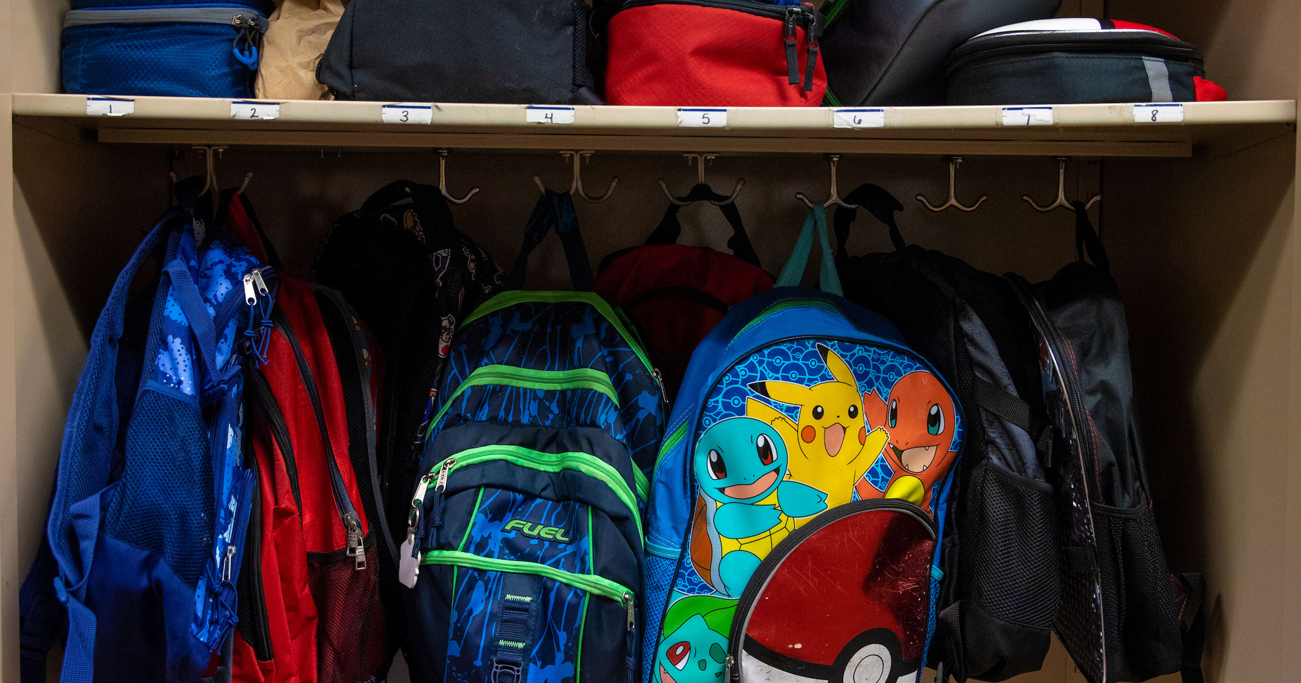 Children’s backpacks hang on numbered hooks in a school storage area beneath a shelf holding lunch bags and other bags.