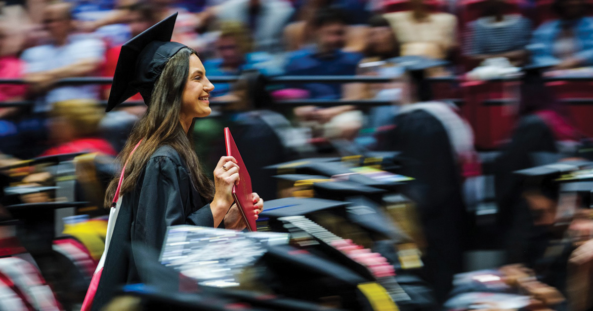 Smiling graduate carries a red diploma cover while walking through the commencement ceremony.