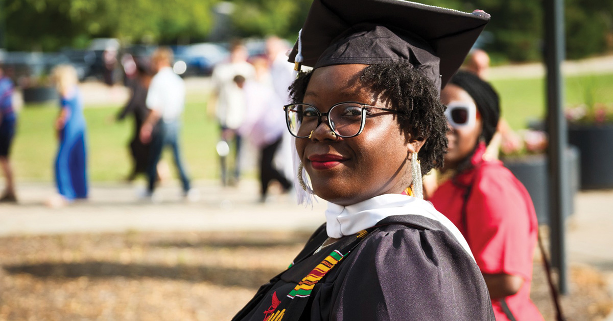 Graduate in cap and gown smiles at the camera during an outdoor commencement celebration.